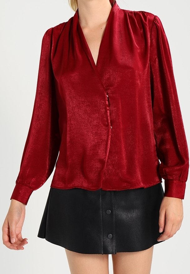 Femme portant un chemisier rouge brillant à manches longues avec un col en V et une jupe en cuir noire boutonnée, debout devant un fond uni.