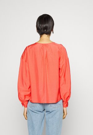 Blouse en coton corail avec de longues manches bouffantes, encolure froncée et coupe floue ; associée à un jean en denim bleu clair.
