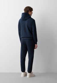 Felpa blu navy con cappuccio regolabile e joggers coordinati. Tessuto liscio, polsini aderenti e dettaglio con tasca posteriore sui pantaloni. Sneakers crema.