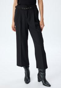 Pantalons larges noirs sur mesure avec une ceinture à boucle rectangulaire, un devant plissé et une texture lisse. Associés à des bottines noires à talons.