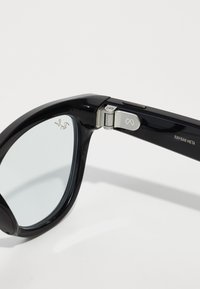 Occhiali da sole neri in plastica con una finitura lucida. Presentano una cerniera in metallo e il logo Ray-Ban sulla tempia. Le lenti sono leggermente oscurate e rotonde.