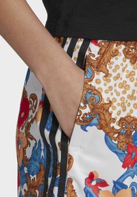 Pantalones cortos de colores variados con intrincados diseños en dorado y floral, franjas laterales negras y una mano en el bolsillo lateral.