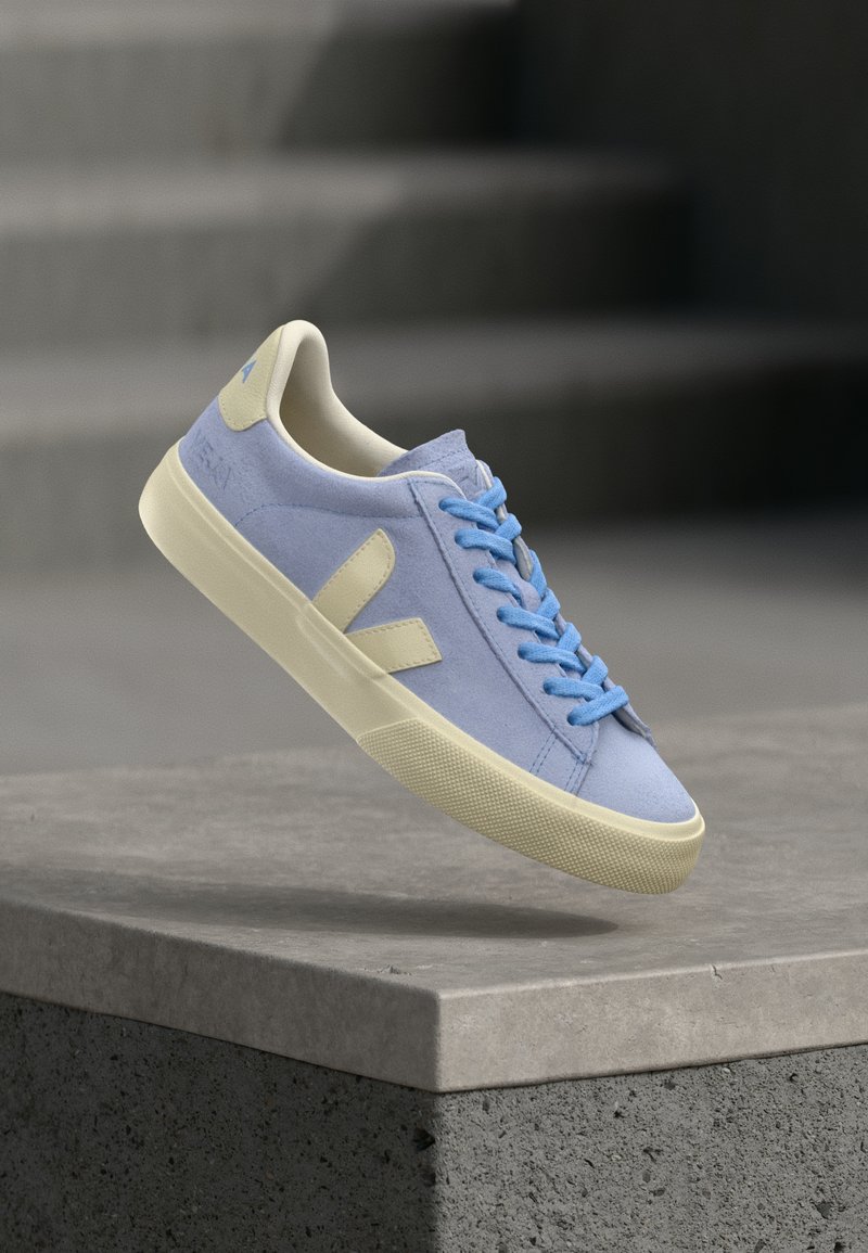 Ανοιχτό μπλε σουέντ αθλητικό παπούτσι με κρεμ σόλα και λογότυπο, μπλε κορδόνια, αιωρούμενο πάνω από γκρίζα πέτρινη επιφάνεια με θολωμένες σκάλες στο φόντο.