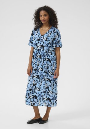 Maxi dress in light blue fabric featuring a floral pattern in black and darker blue. V-neckline, short sleeves, and cinched waist. Black flat shoes.