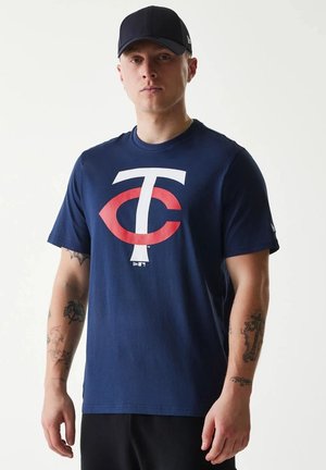 Jeune homme portant une casquette de baseball bleu marine et un T-shirt bleu marine avec un grand logo "TC" rouge et blanc, debout devant un fond clair uni.