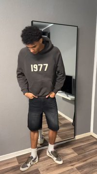 Jeune homme aux cheveux bouclés portant un sweat à capuche sombre marqué "1977", un short noir, des chaussettes blanches et des baskets grises, debout devant un miroir en pied.