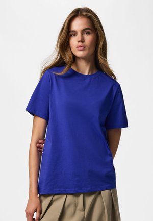 Młoda kobieta z długimi włosami, ubrana w gładki niebieski t-shirt z krótkim rękawem i beżowe plisowane spodnie, stojąca na jednolitym tle.