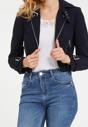 Femme portant une veste courte noire avec des fermetures éclair et des boucles argentées, un top blanc bordé de dentelle et un jean bleu taille haute.