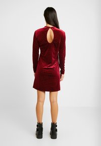 Sammet, burgundy klänning med långa ärmar, hög krage och nyckelhålsrygg. Dekorerad med små vita prickar, kombinerad med svarta ankelstövlar.