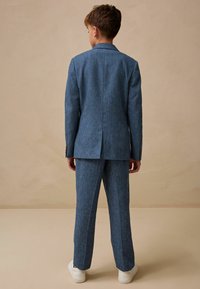 Completo blu testurizzato con un design slim fit, caratterizzato da una singola fessura sul retro, abbinato a sneakers bianche per un look contemporaneo.
