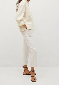 Jasno beżowa koszula w oversize z dwiema kieszeniami na froncie, w połączeniu z dopasowanymi białymi spodniami i brązowymi sandałami na paski. Gładki, elegancki design.