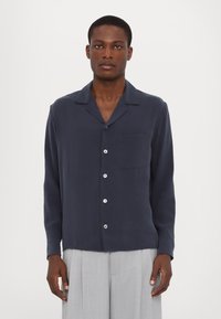 Dunkelblaues Langarmhemd mit Kragen, leichter Knopfleiste und Brusttasche. Weicher Stoff, taillierte Passform, kombiniert mit hellgrauen Hosen.