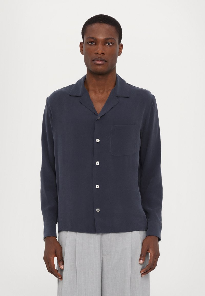 Camisa de manga larga azul marino con cuello, botonadura ligera en el frente y un bolsillo en el pecho. Tejido suave, ajuste entallado, combinada con pantalones gris claro.