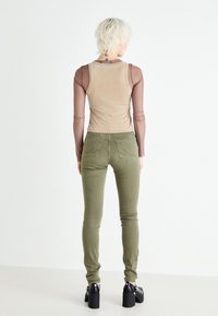 Top de malla de manga larga en marrón con una camiseta beige debajo, combinado con jeans ajustados verdes oliva. Unos zapatos de tacón grueso negros completan el look.
