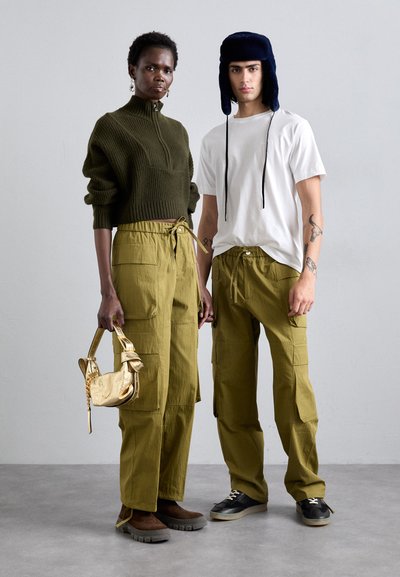 Two models wear casual outfits: a green knit sweater, white tee, and olive cargo pants. Accessories include a shiny gold bag and navy hat.