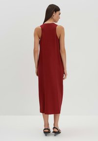 Bordeauxfarbenes, ärmelloses Maxikleid mit lockerem Schnitt, glatter Textur und seitlichem Schlitz für Bewegungsfreiheit, kombiniert mit schwarzen Absatzsandalen.