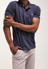 Homme portant un polo bleu marine à manches courtes et un pantalon beige, une main dans la poche et l'autre tenant son bras opposé.