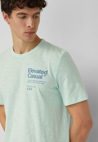Mintgroen T-shirt met korte mouwen en een ronde hals. Voorzijde met blauwe bedrukt tekst: "Elevated Casual" en merkinformatie. Zachte, gestructureerde stof.