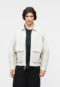 Νεαρό άτομο με μεσαίου μήκους σκούρα μαλλιά φοράει λευκό μπουφάν με φερμουάρ πάνω από μαύρο πουκάμισο και μαύρο παντελόνι, στέκεται μπροστά σε απλό φόντο.