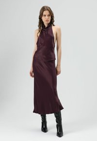 Bordeauxrode satijnen halterjurk met een vloeiende rok en een draperiehals. Gecombineerd met kniehoge zwarte puntige laarzen.