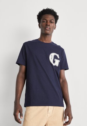 Camiseta de algodón azul marino con escote redondo, mangas cortas y un logo circular blanco que presenta "GANT" y una "G" en el pecho.