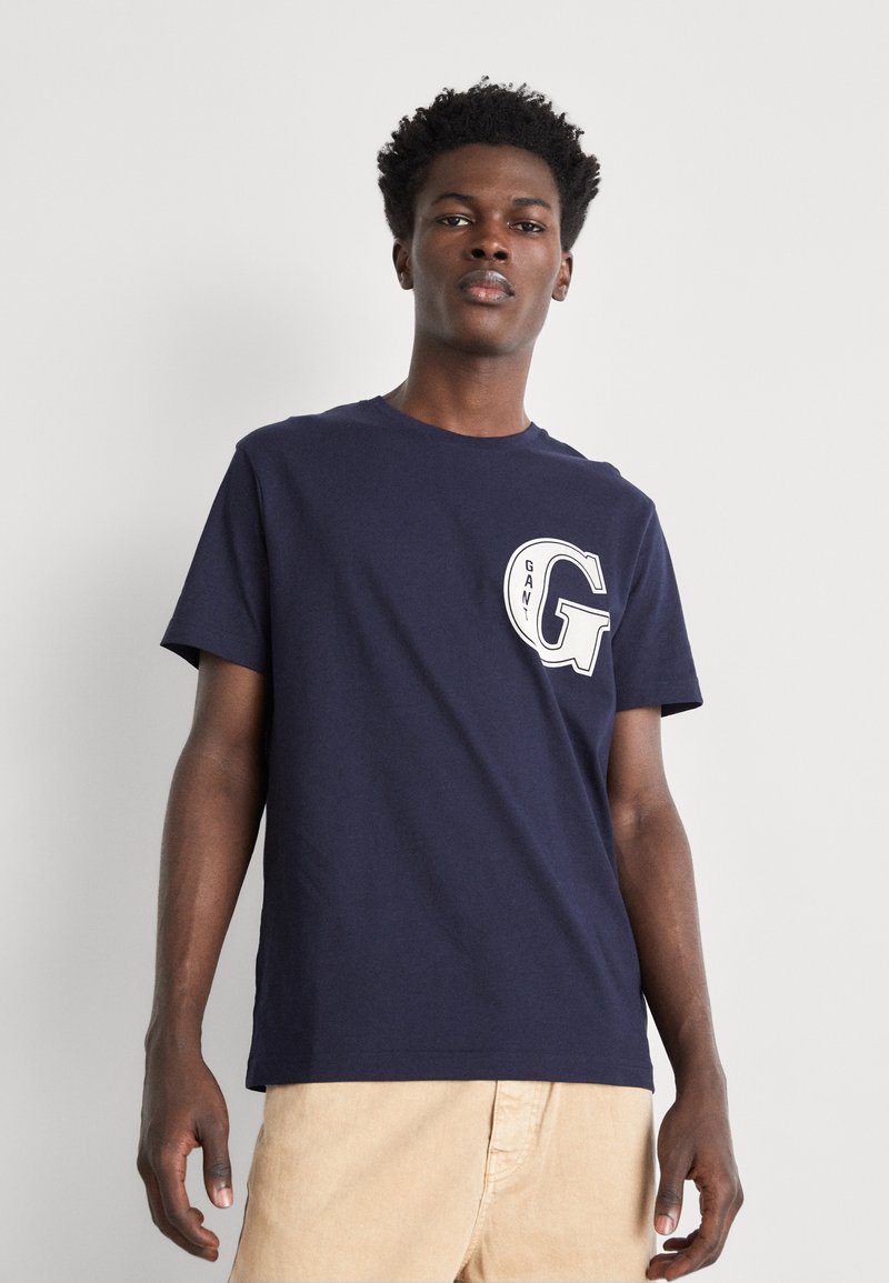 Camiseta de algodón azul marino con escote redondo, mangas cortas y un logo circular blanco que presenta "GANT" y una "G" en el pecho.