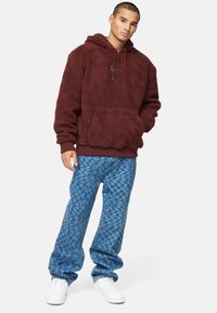 Felpa Bordeaux con cappuccio e tasca frontale; abbinata a jeans blu a fantasia e sneakers bianche. Vestibilità comoda e materiali testurizzati.