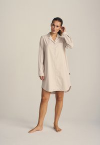 Camicia da notte con bottoni di un leggero beige, caratterizzata da righe verticali e una tasca sul petto, realizzata in tessuto morbido con maniche lunghe e orlo arrotondato.