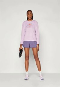 Ανοιχτό μωβ μακρυμάνικη μπλούζα με γραφικό βουνού, συνδυασμένη με σκούρες μωβ βερμούδες. Το μοντέλο κρατάει ένα μαύρο μπουκάλι νερού και φοράει ανοιχτά αθλητικά παπούτσια.