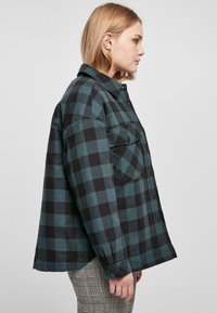 Camicia a quadri verde e nera, design con bottoni, ampie tasche sul petto e vestibilità relaxed. Il tessuto appare morbido e testurizzato.