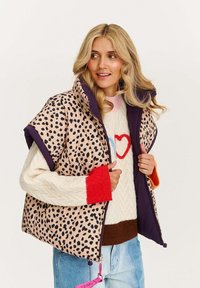 Beżowa kamizelka puff z czarnym printem w panterkę, z wysokim kołnierzem i fioletowymi wykończeniami. Nosi się ją na dzianinowym swetrze w kolorze kremowym z kolorowymi rękawami.
