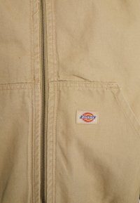 Beige plátěná bunda s centrálním kovovým zipem a přední kapsou, na které je malá obdélníková nášivka s logem Dickies.