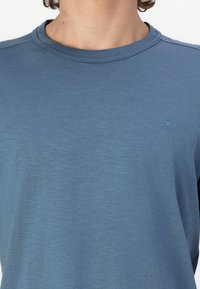 Tricou albastru cu guler rotund, dintr-un material moale și texturat, cu un logo subtil pe piept și mâneci scurte.