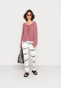 Top de manga longa cor rosa poeira com detalhes texturizados, combinado com calças brancas com padrão de tie-dye preto. Acessorizado com uma bolsa preta.