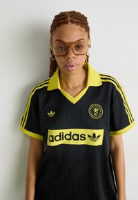 Melns futbola krekls ar dzelteniem akcentiem, svītrainu tekstūru un lielu adidas logo. Apkakle ir dzeltena, ar trim svītrām uz piedurknēm.