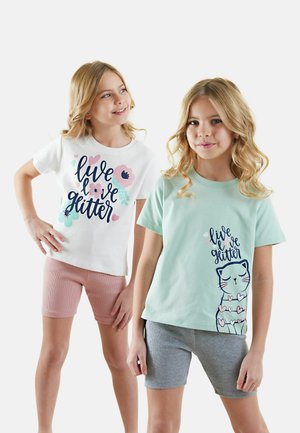 Kaksi nuorta tyttöä wear t-paitoja, toinen valkoinen, jossa on kukkakuosi ja teksti, toinen mintunvihreä kissan kuvalla. Molemmilla on shortsit.