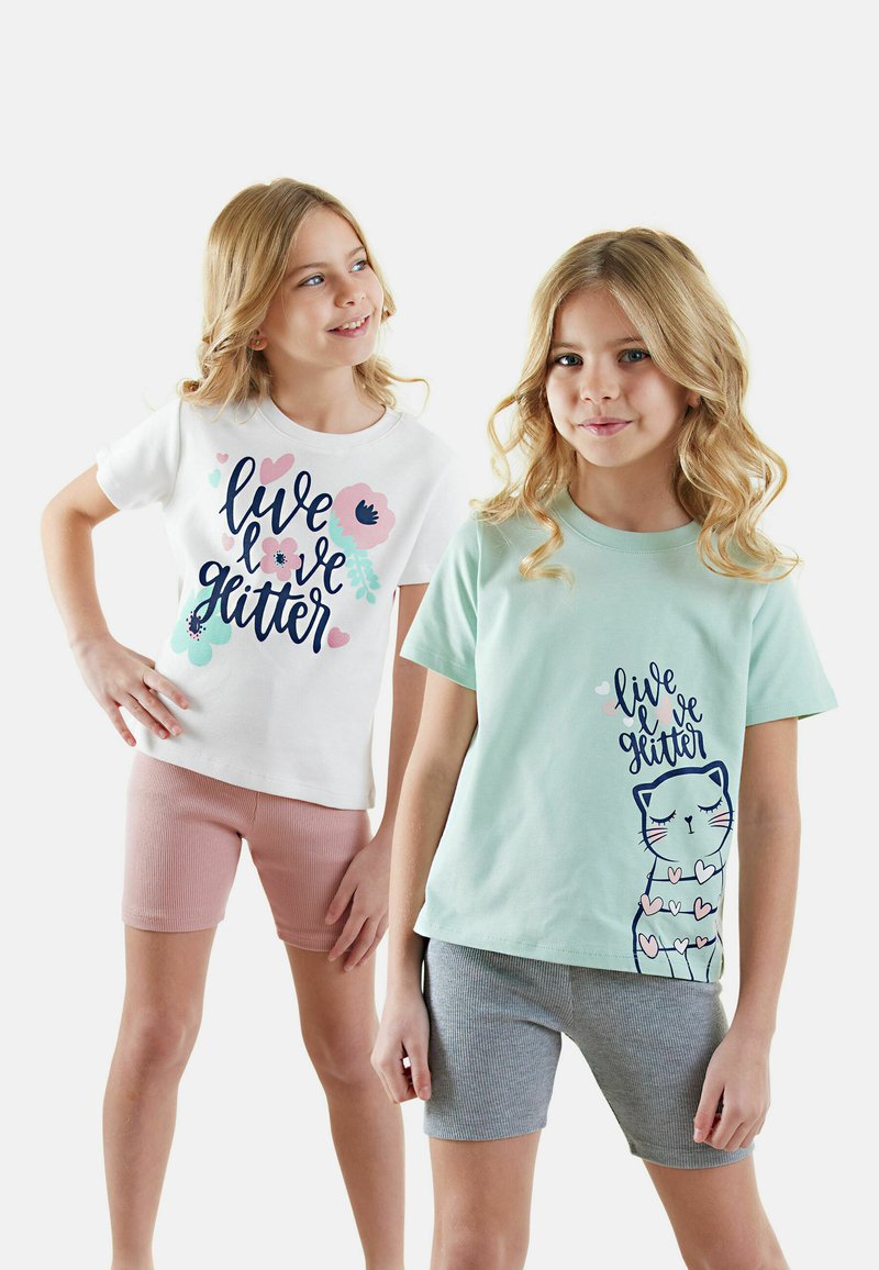 Zwei junge Mädchen tragen T-Shirts, eines weiß mit einem floralen Design und Text, das andere mintgrün mit einer Katzenillustration. Beide tragen Shorts.