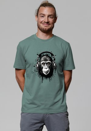 Mann lächelt und trägt ein grünes T-Shirt mit einer detaillierten Grafik eines Schimpansengesichts, das Kopfhörer trägt.