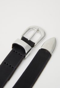 Cinturón de cuero negro con hebilla de metal plateado y punta de metal afilada sobre un fondo blanco.