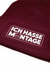 Maroon knitted beanie featuring a rectangular patch with the text "ICH HASSE MONTAGE" and a cat graphic in white.