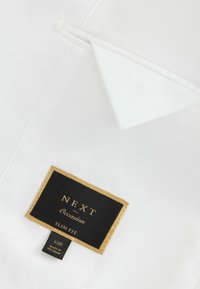 Fehér szövet blézer, slim fit kialakítással, fekete címkével, amely arany betűkkel hirdeti a "NEXT Occasion Slim Fit" feliratot.
