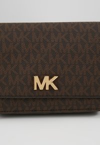 Ruskea teksturoitu lompakko, jossa on toistuvia "MK"-kirjaimia kuvioina ja kultainen "MK"-logoembleemi keskittyen etuoveen.