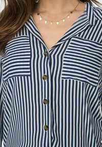 Camisa de botones en rayas azules y blancas, con dos bolsillos en el pecho, un cuello y botones de bronce. Tejido suave con un diseño clásico.