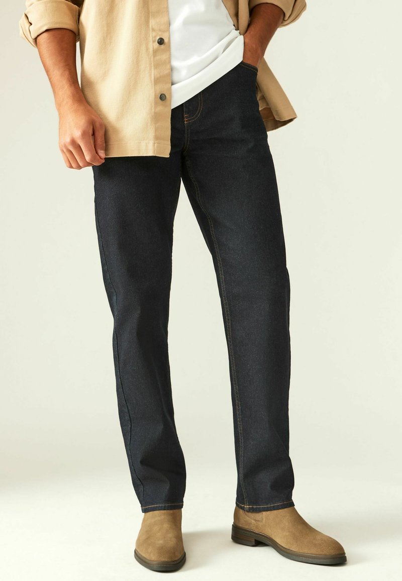 Jeans de mezclilla oscura con un diseño de pierna recta, adornados con costuras naranjas y un ligero brillo, combinados con botas de gamuza en color beige.