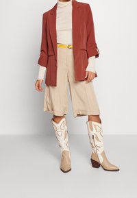 Rostfärgad blazer över en beige ribbad topp, högmidjade beige shorts och vita cowboystövlar med bruna accenter och fjärilsdesign.