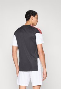 Αθλητική φανέλα σε μαύρο και άσπρο χρώμα με κόκκινες λεπτομέρειες. Κοντά μανίκια, λεία υφή, στρογγυλή λαιμόκοψη και χαρακτηριστικό σχεδιασμό με τρεις ρίγες στους ώμους.