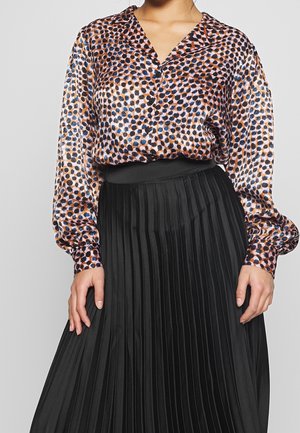Vrouw draagt een blouse met lange mouwen en knopen, versierd met meerkleurige stippen, en een zwarte geplooide hoge taille rok.