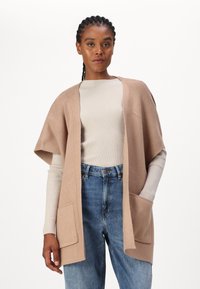 Cardigan beige aperto con maniche corte, texture a maglia e due tasche frontali, indossato sopra un maglione leggero a coste e jeans blu.