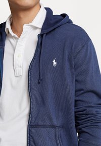 Mörkblå zip hoodie i mjukt material med en vit polo-logotyp på vänster bröst, med huva, framfickor och strukturerad yta.
