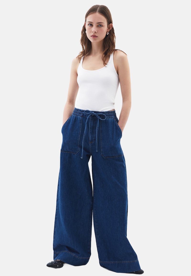 Modré široké džíny s elastickým pasem a dvěma předními kapsami, spárované s přiléhavým bílým topem na ramínka. Hladká textura.